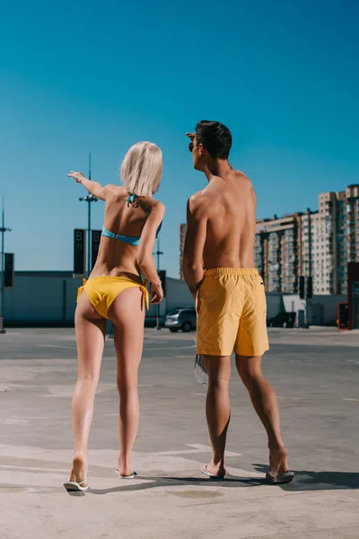 Rear view of young couple in bikini and swimming shorts pointing somewhere on parking — Stock Photo