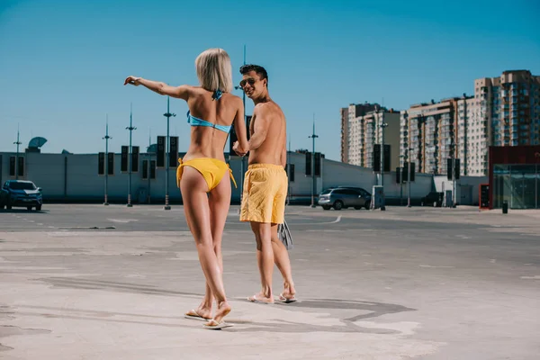 Vista posteriore della giovane coppia attraente in bikini e pantaloncini da bagno che punta da qualche parte sul parcheggio — Foto stock