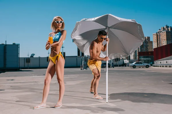 Young attractive couple with umbrella in swimsuits tanning on parking — Stock Photo