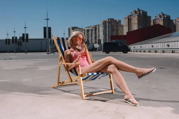 Schöne junge Frau trinkt Cocktail, während sie in der Sonnenliege auf dem Parkplatz sitzt — Stockfoto