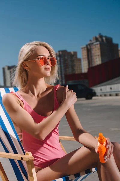 Close-up portrait of young woman in pink swimsuit sitting on sun lounger and applying sunscreen lotion on parking — Stock Photo
