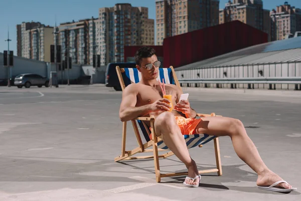 Молодой мужчина без рубашки расслабляется на шезлонге с коктейлем и использует смартфон на парковке — стоковое фото