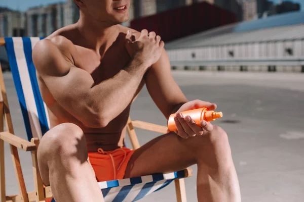 Cropped shot of shirtless man sitting on sun lounger and applying sunscreen lotion on parking — Stock Photo