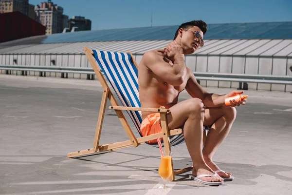 Attractive young shirtless man sitting on sun lounger and applying sunscreen lotion on parking — Stock Photo