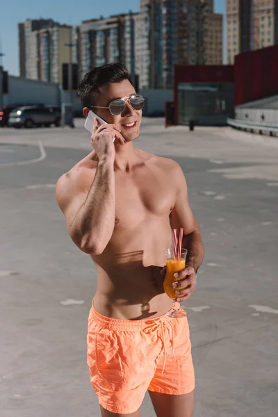 Красивий молодий без сорочки чоловік у плавальних шортах з коктейлем розмовляє по телефону на парковці — стокове фото