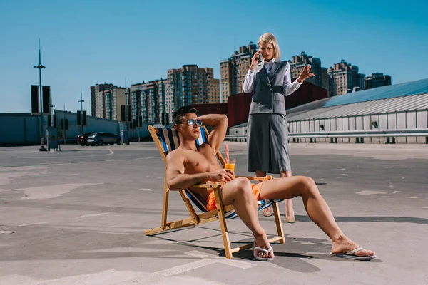 Schöner junger Mann entspannt sich auf einer Sonnenliege, während seine verrückte Chefin im Anzug neben ihm steht und auf dem Parkplatz telefoniert — Stockfoto