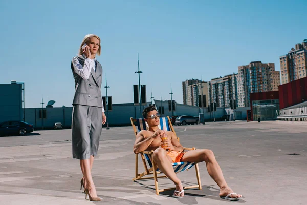 Attractive young man relaxing on sun lounger while his lady boss in suit next to him and talking by phone on parking — Stock Photo