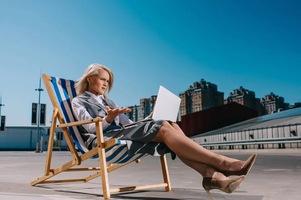 Mad young businesswoman in formal wear sitting in sun lounger and looking at laptop on parking — Stock Photo