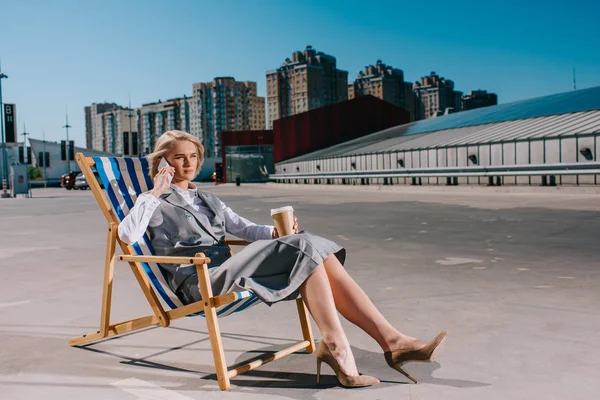 Attraktive junge Geschäftsfrau in formeller Kleidung sitzt in einer Sonnenliege mit Coffee to go und telefoniert auf dem Parkplatz — Stockfoto