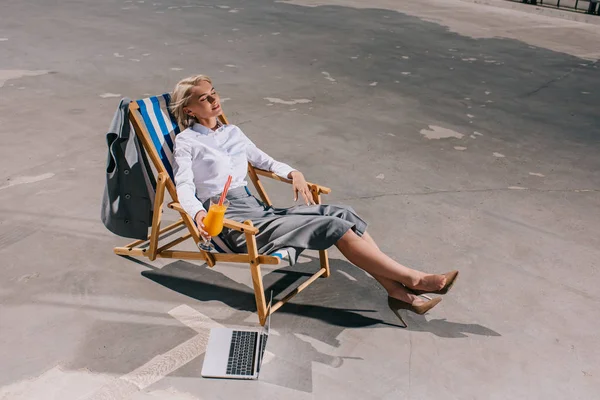 Hochwinkelaufnahme der schönen jungen Geschäftsfrau in formeller Kleidung, die in einer Sonnenliege mit Cocktail auf Asphalt sitzt — Stockfoto