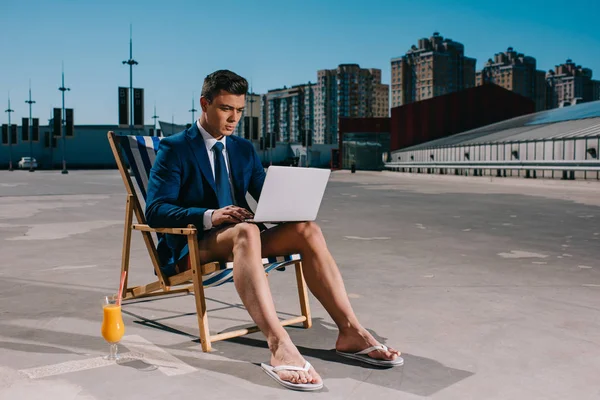 Bonito jovem empresário em shorts e jaqueta trabalhando com laptop enquanto sentado na espreguiçadeira no estacionamento — Fotografia de Stock
