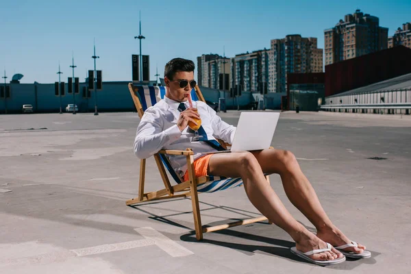 Beau jeune homme d'affaires en short travaillant avec ordinateur portable tout en étant assis sur une chaise longue sur le parking et en buvant un cocktail — Photo de stock