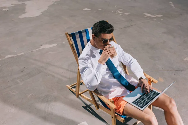 Vue grand angle de jeune homme d'affaires en short travaillant avec ordinateur portable tout en étant assis sur une chaise longue sur de l'asphalte et en buvant du café à emporter — Photo de stock