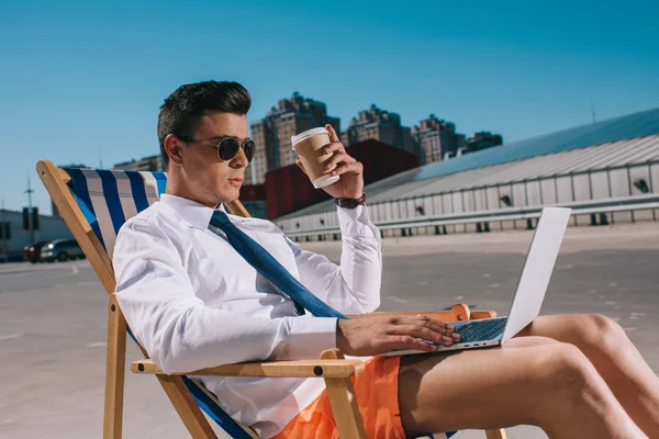 Beau jeune homme d'affaires en short avec café pour aller travailler avec ordinateur portable tout en étant assis sur une chaise longue sur le parking — Photo de stock