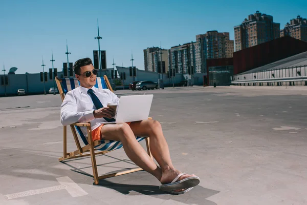 Handsome young businessman in shorts working with laptop while sitting on sun lounger on parking — Stock Photo