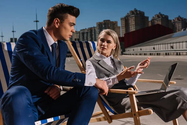 Jeunes gens d'affaires travaillant avec un ordinateur portable tout en étant assis sur des chaises longues sur le parking — Photo de stock