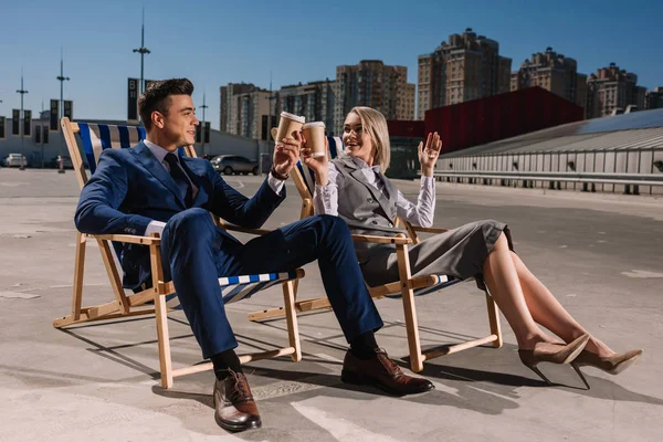 Young happy business people relaxing on sunbeds and clinking glasses with coffee to go on parking — Stock Photo