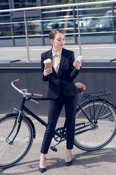 Junge Geschäftsfrau mit Coffee to go und Smartphone steht neben Retro-Fahrrad auf der Straße — Stockfoto