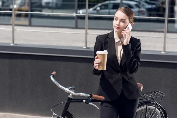 Young businesswoman with coffee to go talking on smartphone while standing near retro bicycle on street — Stock Photo