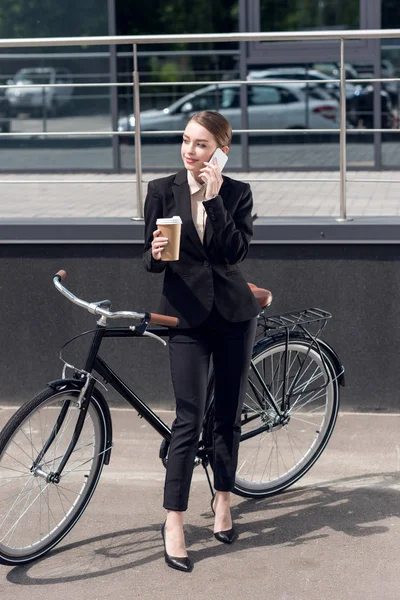 Young businesswoman with coffee to go talking on smartphone while standing near retro bicycle on street — Stock Photo