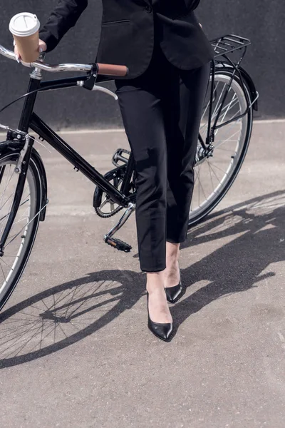 Cropped shot of businesswoman in suit with coffee to go standing near retro bicycle on street — Stock Photo
