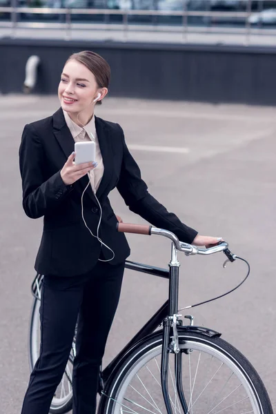 Portrait of smiling businesswoman in earphones with smartphone standing near retro bicycle on street — Stock Photo