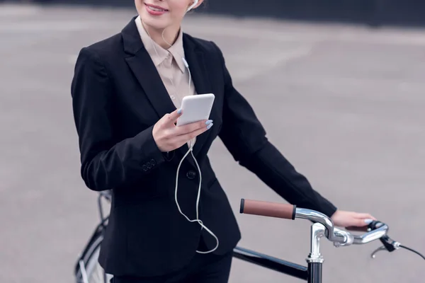Recortado disparo de mujer de negocios en auriculares con teléfono inteligente de pie cerca de bicicleta retro en la calle — Stock Photo