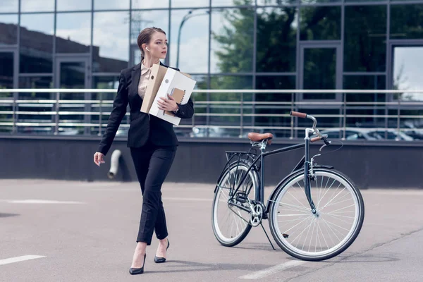 Joven empresaria en auriculares con documentos caminando por la calle con bicicleta aparcada detrás - foto de stock