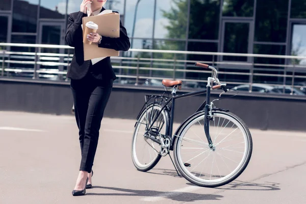 Cropped shot of businesswoman with documents and coffee to go talking on smartphone while walking on street with bicycle parked behind — Stock Photo