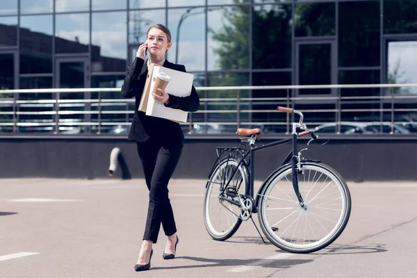 Businesswoman with documents and coffee to go talking on smartphone while walking on street with bicycle parked behind — Stock Photo