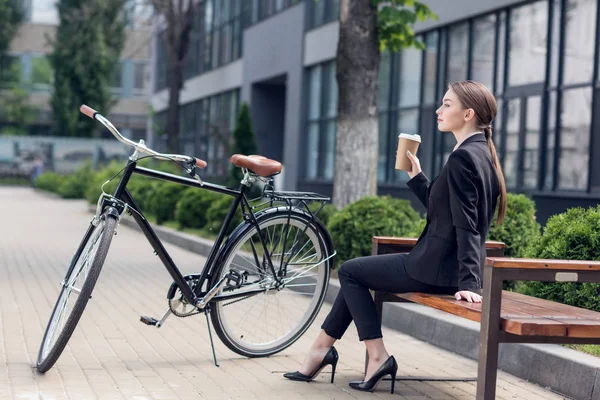 Боковой вид молодой бизнесвумен с кофе, чтобы отправиться отдыхать на скамейку с припаркованным рядом ретро-велосипедом — стоковое фото