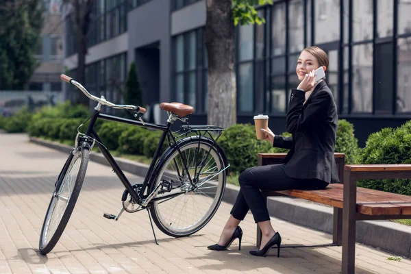 Молодая деловая женщина с кофе, чтобы пойти говорить на смартфоне во время отдыха на скамейке — стоковое фото