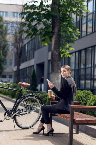 Усміхнена молода бізнес-леді зі смартфоном і кавою, щоб піти відпочивати на лавці — стокове фото