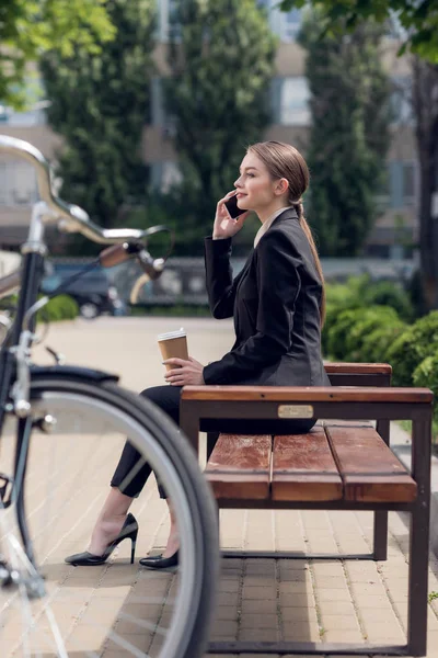 Молода бізнес-леді з кавою, щоб піти говорити по смартфону, відпочиваючи на лавці — стокове фото