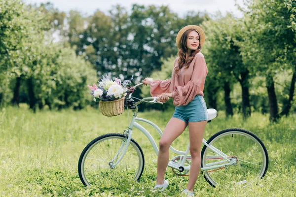 Joven hermosa mujer de pie cerca de bicicleta retro con canasta de mimbre llena de flores en el bosque - foto de stock