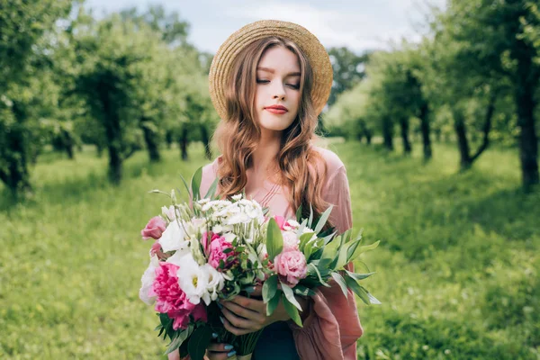 Portrait of young woman in hat with bouquet of flowers in hands in park — Stock Photo