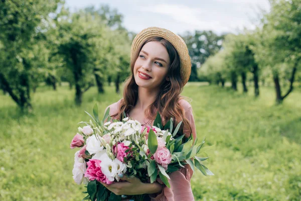 Portrait of beautiful smiling woman in hat with bouquet of flowers looking away in park — Stock Photo