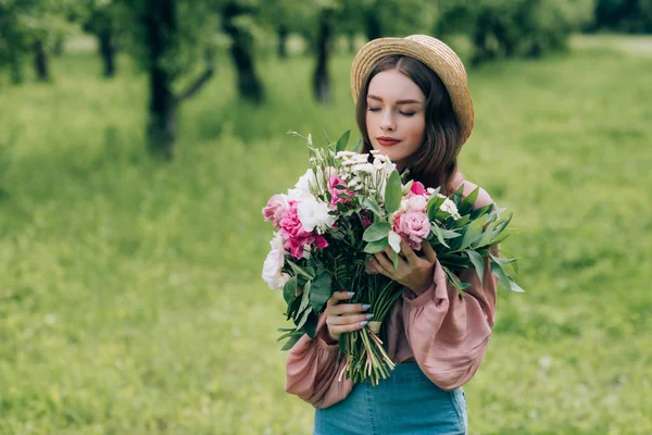 Portrait of beautiful pensive woman in hat with bouquet of flowers in park — Stock Photo