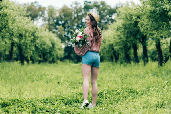 Vista posteriore di donna sorridente in cappello con mazzo di fiori in mano in piedi nel parco — Foto stock