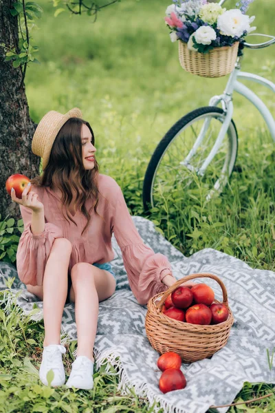 Jolie femme souriante en chapeau reposant sur une couverture avec panier en osier avec pommes et vélo garé à proximité dans le parc — Photo de stock