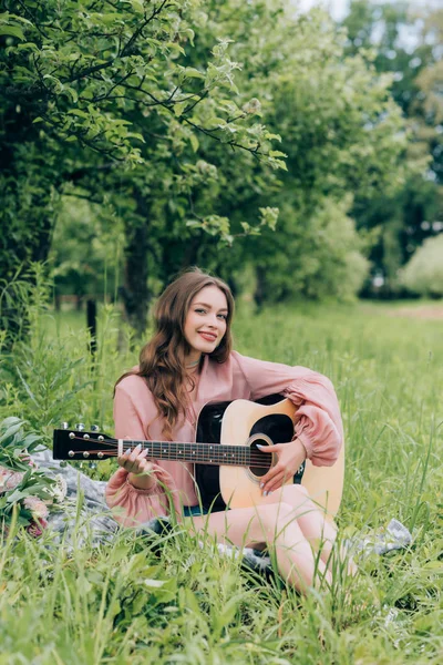 Молода усміхнена жінка з акустичною гітарою відпочиває на ковдрі з букетом квітів у парку — стокове фото
