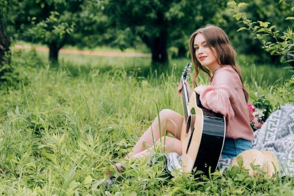 Vue latérale de jeune femme avec guitare acoustique reposant sur une couverture avec bouquet de fleurs dans le parc — Photo de stock