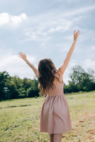 Rear view of woman in stylish dress with outstretched arms standing in meadow with blue sky on background — Stock Photo
