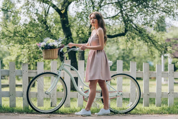 Вид збоку молодої жінки в одязі з ретро велосипедом з плетеним кошиком, повним квітів у сільській місцевості — стокове фото