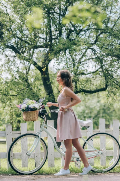 Vista lateral da jovem mulher de vestido com bicicleta retro com cesta de vime cheia de flores no campo — Fotografia de Stock