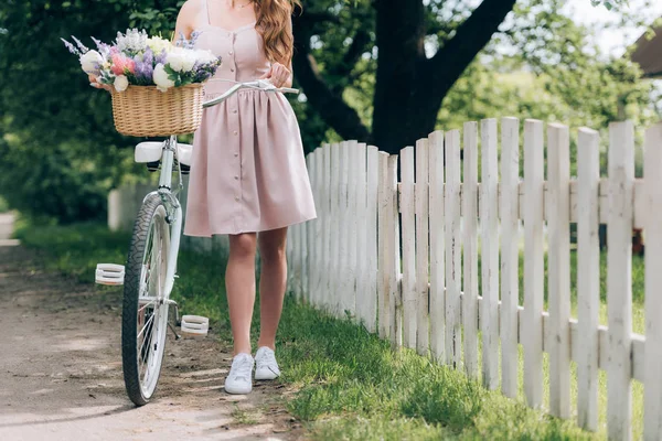 Vista parcial de mujer en vestido con bicicleta retro con cesta de mimbre llena de flores en el campo - foto de stock