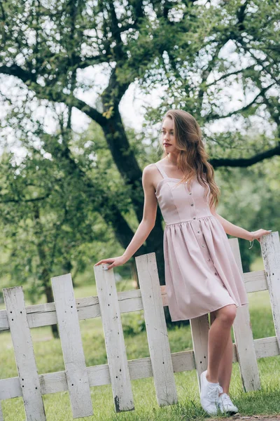 Красива вдумлива жінка в стильній сукні, що стоїть на білому паркані в сільській місцевості — стокове фото