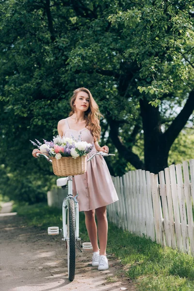 Beautiful pensive woman in dress with retro bicycle with wicker basket full of flowers at countryside — Stock Photo