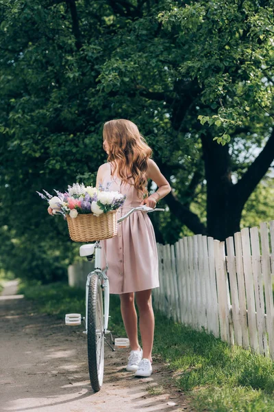 Giovane donna in abito con bicicletta retrò con cesto di vimini pieno di fiori in campagna — Foto stock