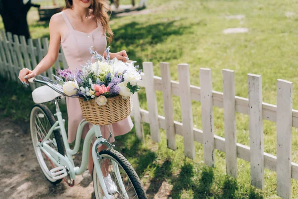 Vista parcial de la mujer en vestido elegante con bicicleta retro con cesta de mimbre llena de flores en el campo - foto de stock